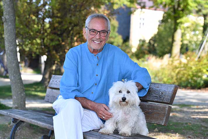 Der Kunstmaler Franco Thamér sitzt mit seinem Hund Lui auf einer Parkbank am Chiemsee
